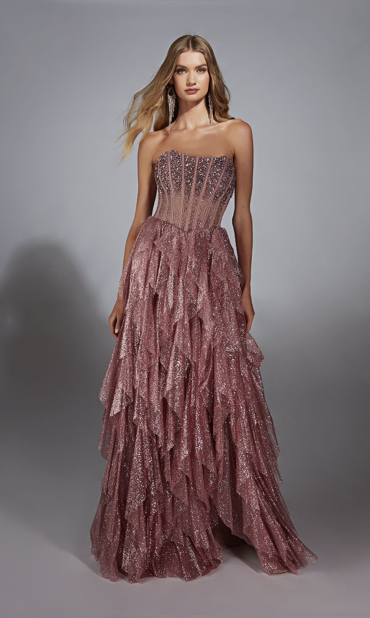 A woman poses against a gray background wearing the Strapless Long Glitter A-Prom Dress: Alyce 61762, a pink, ruffled evening gown.